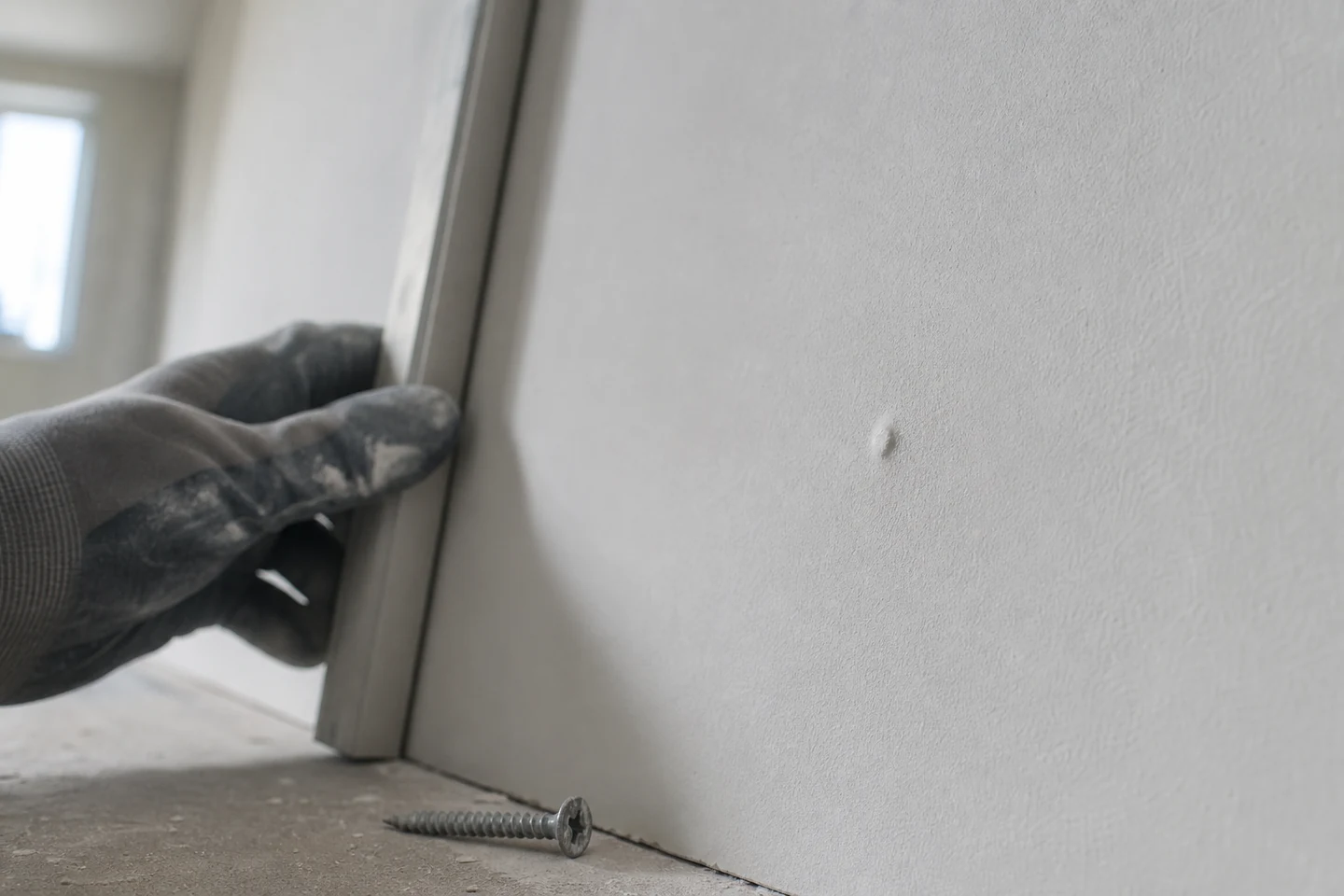 Pared de pladur terminada con iluminación lateral que muestra un pequeño tornillo salido junto a un técnico que revisa la superficie con una regla.